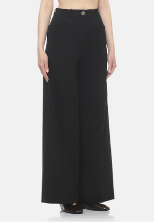 Donna che indossa pantaloni neri a vita alta e gamba larga con chiusura a bottone e cerniera, in piedi con le braccia rilassate ai lati.
