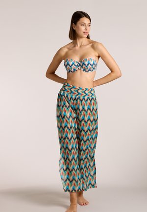 Frau steht barfuß und trägt ein trägerloses, buntes Oberteil mit Zickzackmuster sowie passende weit geschnittene Hosen vor einfarbigem Hintergrund.