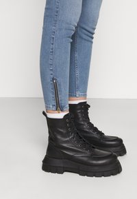 Botas de combate de cuero negro con suelas gruesas, con cordones y un diseño sin costuras; llevadas con jeans ajustados azules que tienen cremalleras laterales.