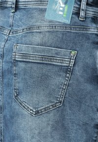 Jean azul de mezclilla con un solo bolsillo trasero que presenta detalles de costura y una etiqueta de tela adjunta con un acento verde. Textura ligeramente descolorida.