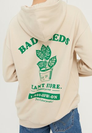 Sweat à capuche beige avec un graphisme vert de plante en pot et le texte « BAD SEEDS PLANT STORE 1-800-GROW-ON » dans le dos, porté avec un jean bleu.
