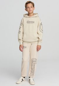 Beżowa bluza z kapturem z czarnym nadrukiem tekstu z przodu i na rękawach, w zestawie z dopasowanymi beżowymi dresami z nadrukiem logo, noszone z białymi trampkami.