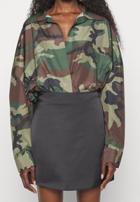 Donna che indossa una camicia a maniche lunghe in camouflage infilata in una gonna nera a vita alta, ripresa dalle spalle fino a metà coscia.