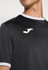 Schwarzes Sportshirt aus glattem Stoff, mit einem weißen Kragen und einem weißen Logo im oberen linken Brustbereich.