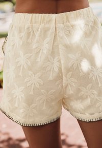 Pantaloni corti color crema realizzati in tessuto testurizzato, con un motivo di palme e cuciture a contrasto lungo l'orlo. Dettaglio della vita elasticizzata.