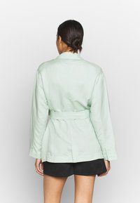 Veste en lin vert clair avec une taille cintrée, des manches longues et un design ceinturé. Présente des détails de boutons sur les poignets.
