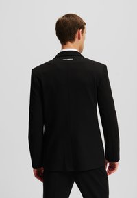 Schwarzer maßgeschneiderter Blazer mit klassischem Kragen, strukturierter Stoff, einziger Schlitz auf der Rückseite und kleinem weißen Logo am Halsausschnitt.
