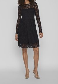 Black lace dress with long sleeves, fitted bodice, and flared skirt. Scalloped hem detail and textured overlay. Worn with gold heels.