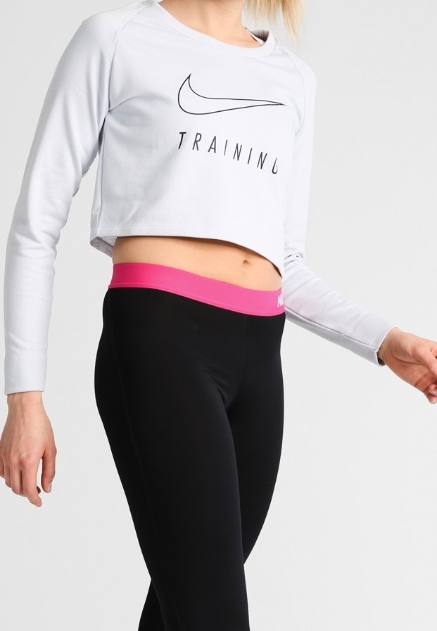 Haut d'entraînement blanc court avec un logo noir, associé à un legging noir avec une taille rose. Tissu lisse, design ajusté.