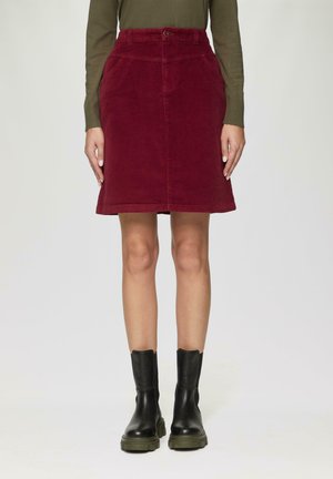 Jupe en velours côtelé bordeaux avec une taille ajustée, des poches latérales et un ourlet évasé. Associée à des bottines noires à semelles épaisses pour un contraste.