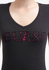 Černé tričko s výstřihem do V, zdobené výrazným nápisem "GUESS" v růžovém zvířecím potisku s texturovaným povrchem.