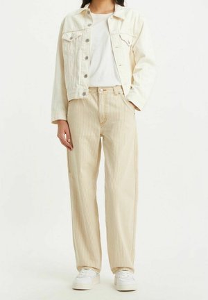 Kobieta w kremowej kurtce denimowej, białym t-shircie, beżowych paskowanych szerokich spodniach i białych trampkach, stoi z jedną ręką w kieszeni.