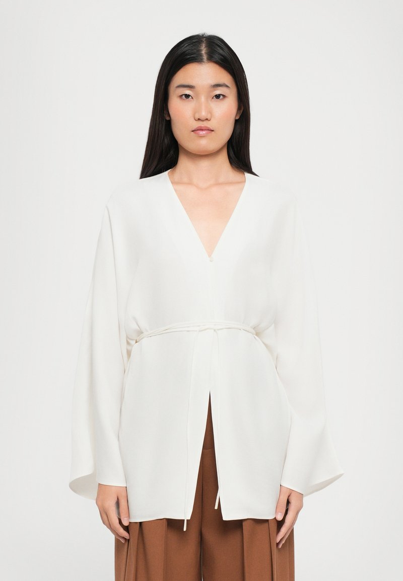 Woman with long dark hair wearing a white V-neck blouse with wide sleeves and a tied waist, paired with brown pleated pants.