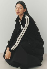 Veste noire avec des rayures blanches athlétiques sur les manches, fermeture éclair, col haut et coupe décontractée. Le tissu semble lisse et texturé.