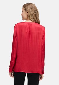 Blouse en soie rouge à coupe ample, manches longues et détail de pli subtil à l'arrière. Texture lisse avec un léger éclat.