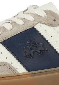 Vita lädersneaker med marinblå mockadetaljer, texturerad marinblå lapp med en logotyp och gummiyttersula. Snörningarna är vita.