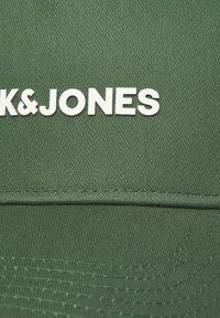 Grøn kasket lavet af tekstureret stof med hvid "K&JONES" logo. Designet inkluderer syede detaljer og en buet skygge.