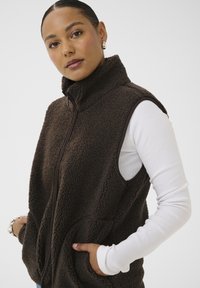 Mujer con un chaleco de forro polar sin mangas de color marrón oscuro sobre una camisa blanca de manga larga, con las manos en los bolsillos del chaleco, mirando hacia la cámara.