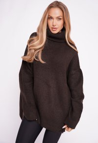 Dunkelbrauner, oversized Strickpullover mit hohem Rollkragen, gerippter Textur, überschnittenen Schultern und seitlichen Schlitzen, kombiniert mit schwarzen Leggings.