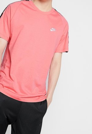 Person trägt ein pinkes Nike-T-Shirt mit schwarzen Hosen und steht mit einer Hand in der Tasche vor einem schlichten hellen Hintergrund.