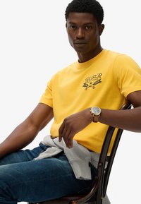 Camiseta de algodón amarilla con el logo "SUPER", combinada con vaqueros azul oscuro y un sudadera gris atada a la cintura; reloj dorado en la muñeca.