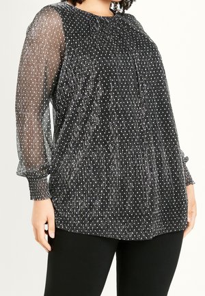 Black sheer long-sleeve blouse with white polka dots worn with black pants, showing textured fabric and cuffed semi-transparent sleeves.