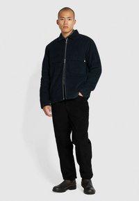 Veste en velours côtelé bleu marine avec col, fermeture éclair à l'avant et deux poches à rabat ; assortie à un pantalon en velours côtelé noir et des chaussures en cuir marron.