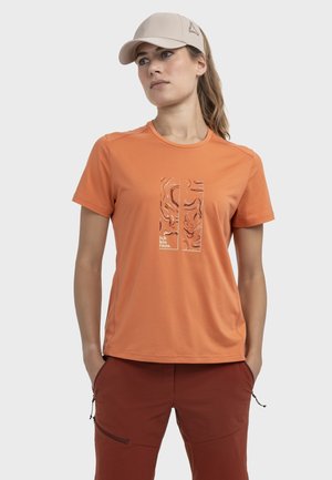 Frau trägt beige Mütze, orangefarbenes kurzärmeliges Shirt mit abstraktem Design und dem Text "Ich bin raus" sowie rostfarbene Hosen mit Reißverschlusstaschen.