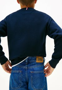Mørkeblå cropped sweatshirt med ribbede manchetter og en rød og hvid stribet krave, parret med blå jeans med et brunt TOMMY HILFIGER mærke.