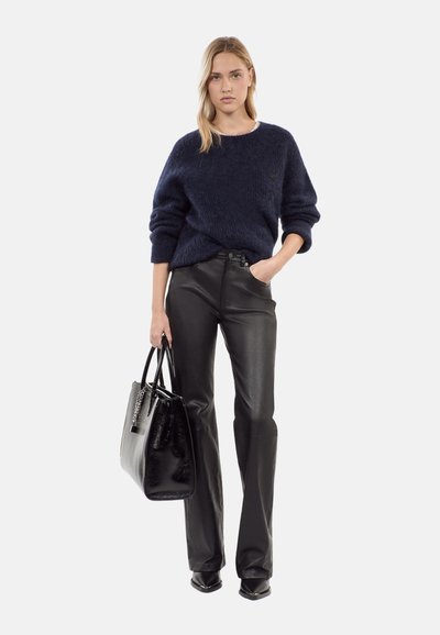 Maglione a maglia blu navy, vestibilità comoda; pantaloni in pelle nera a campana; borsa nera con dettagli in catena; la modella posa con una mano sulla vita.