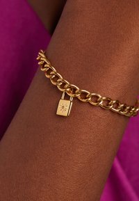 Gouden schakelarmband met grote schakels en een sluiting met een decoratieve hangslot charme, met een gepolijste afwerking en een reflecterend oppervlak.