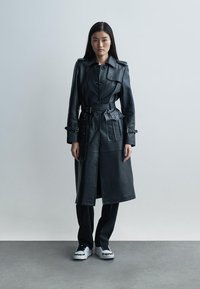 Juodas odinis tranšėjos paltas su dideliais mygtukais, diržu liemens srityje ir šoninėmis kišenėmis; derinamas su juodomis kelnėmis ir baltais sportbačiais.