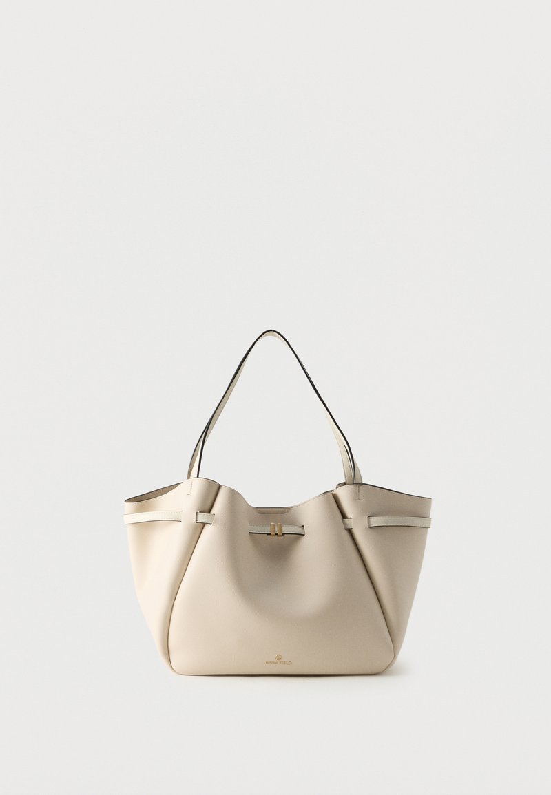 Sac cabas en cuir beige avec deux bretelles et un petit fermoir doré à l'avant, présenté sur un fond blanc uni.