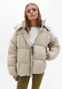 Beige pufferjack met een boxy ontwerp, zichtbare gestikte secties, een hoge kraag en ritssluiting. Gecombineerd met olijfkleurige ribleggings.