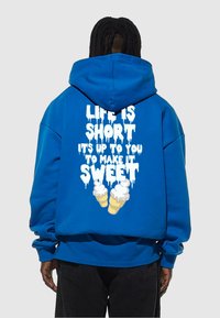 Blå fleecehoodie med en stor vit grafik som säger "LIVET ÄR KORT, DET ÄR UPP TILL DIG ATT GÖRA DET SÖTT," med två glasskoner.