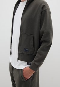 Giacca a zip verde oliva scuro realizzata in tessuto morbido, con cappuccio, tasche laterali e polsini a coste. Indossata sopra una maglietta bianca.