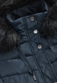 Nærbillede af en mørkeblå quilted vinterjakke med sort faux fur kant, metal tryknapper og blå snore med sølvspidser.
