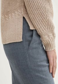 Beżowy sweter dziany z prążkowanymi mankietami i bocznymi rozcięciami, zestawiony z szarymi spodniami o teksturowanej powierzchni. Sweter ma wzór w warkocz.