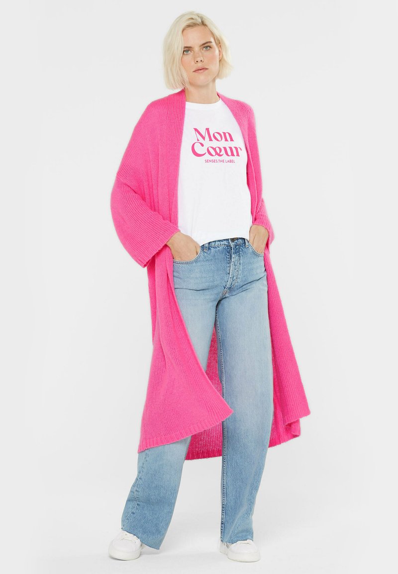 Blonde Frau trägt einen leuchtend pinken langen Cardigan, ein weißes T-Shirt mit pinkem Text, hellblaue Jeans und weiße Sneaker, mit den Händen in den Taschen.