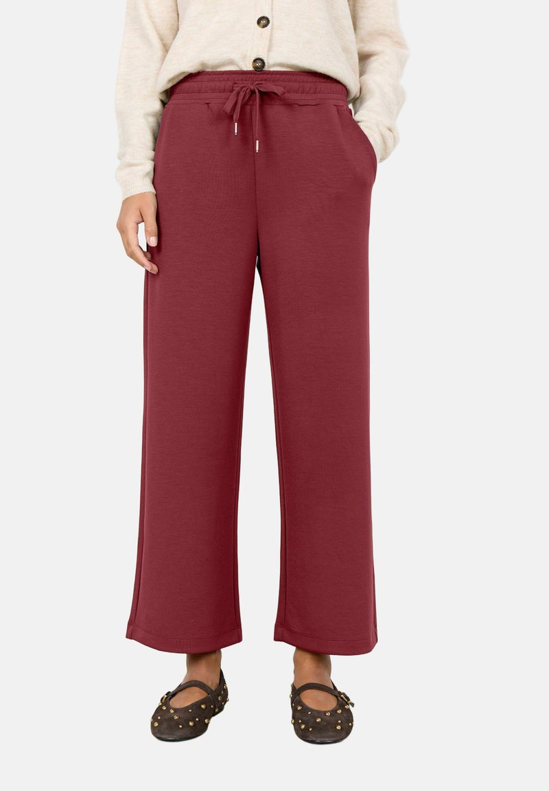 Personne portant un pantalon bordeaux taille haute à jambes larges avec cordon à la taille, un pull beige boutonné et des chaussures Mary Jane marron foncé cloutées.