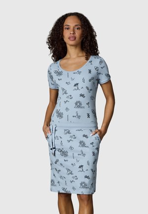 Donna che indossa un vestito azzurro chiaro a maniche corte con disegni lineari neri a tema tropicale e mare, mani nelle tasche della gonna, sfondo neutro.