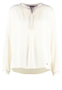Blouse blanche à manches longues avec un col rond, un lien à nouer sur le devant, de subtils plis et des détails de couture noirs sur les poignets et le col.