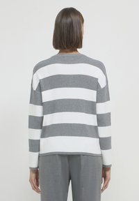 Donna con capelli lisci di lunghezza shoulder che indossa un maglione a maniche lunghe a righe orizzontali grigio e bianco e pantaloni grigi, in piedi con la schiena rivolta.
