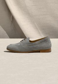 Ανδρικό μοκασίνι από ανοιχτό γκρι σουέτ με λεπτομέρεια κορδονιού, ξύλινη σόλα και διακριτικό ανάγλυφο λογότυπο στο πλάι, εκτεθειμένο σε μπεζ επιφάνεια.