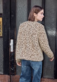 Femme aux cheveux courts bruns portant un pull beige à imprimé léopard et un jean bleu, tendant la main vers la poignée d'une porte vitrée.
