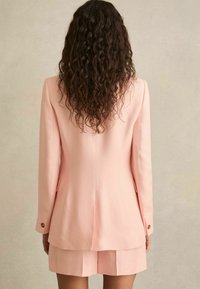 Růžový blazer a kraťasy, s strukturovaným designem, hladkou texturou a knoflíkovými detaily. Pohled zezadu, který ukazuje precizní střih.