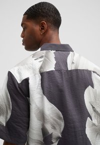 Koszula z krótkim rękawem z dużym abstrakcyjnym wzorem w szaro-białych odcieniach, wykonana z teksturowanej tkaniny, o luźnym kroju i tradycyjnym kołnierzyku.