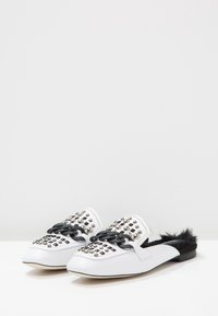 Mules blancs plats avec des embellissements cloutés et une chaîne noire décorative. Doublure en fourrure noire et design à bout pointu.
