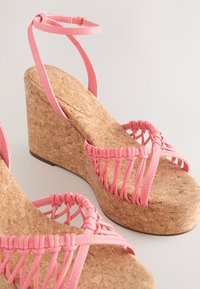 Lyserøde kilehælssandaler med såler af kork, der har flettede stropper og en tynd ankelrem. Glat tekstur med en kontrast af materialer.