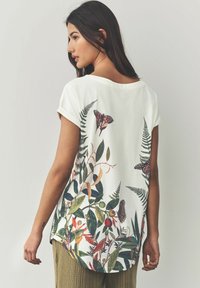 White short-sleeve t-shirt featuring colorful botanical and butterfly prints on the back, with a rounded hem and relaxed silhouette.